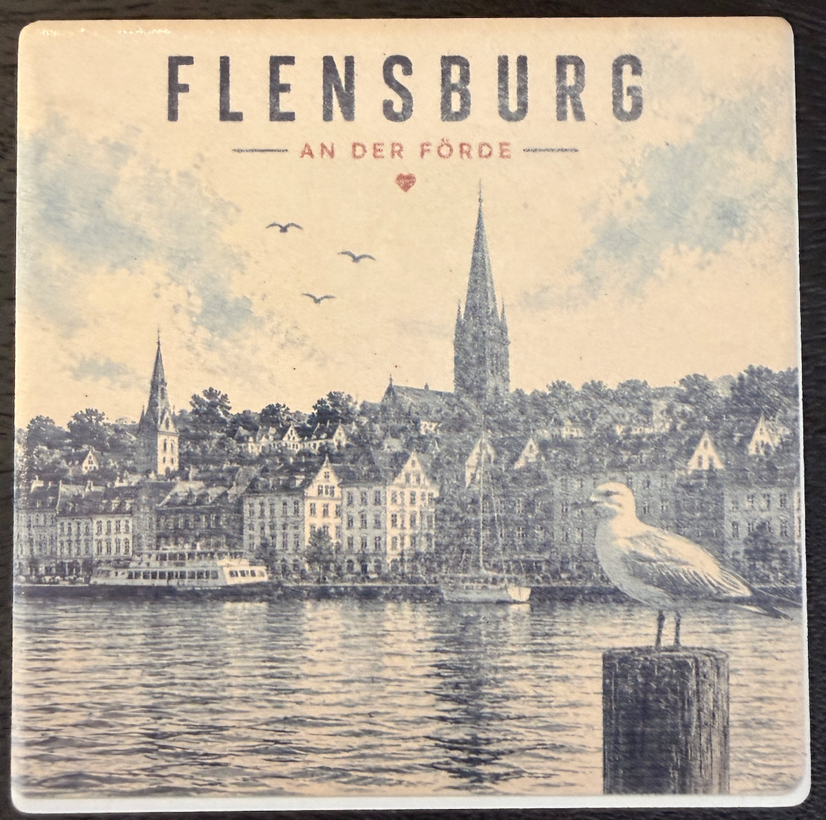 Bedruckte Fliese oder Untersetzer mit einem Vintage-Panorama der Flensburger Förde, Möwe auf einem Dalben und dem Schriftzug „Flensburg an der Förde“.