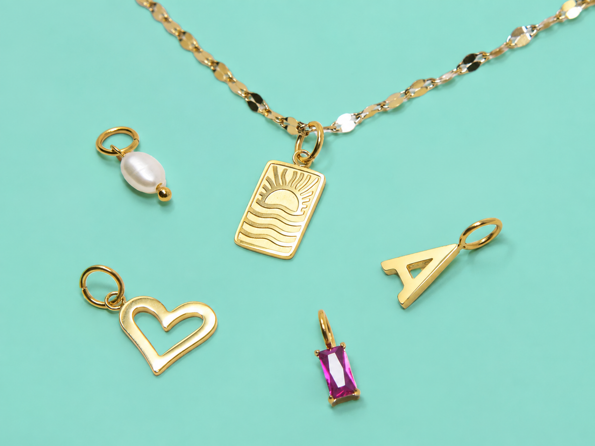 Goldene LJS Schmuckkette mit Sonne-Wellen-Anhänger, Herz, Buchstaben-Charm, Perle und pinkem Schmuckstein auf türkisfarbenem Hintergrund