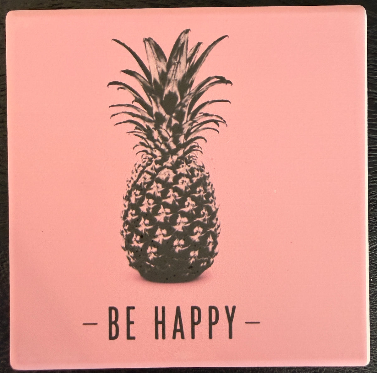 Bedruckte Fliese oder Untersetzer mit einer schwarzen Ananas auf rosafarbenem Hintergrund und dem Schriftzug „Be Happy“ im minimalistischen Design.