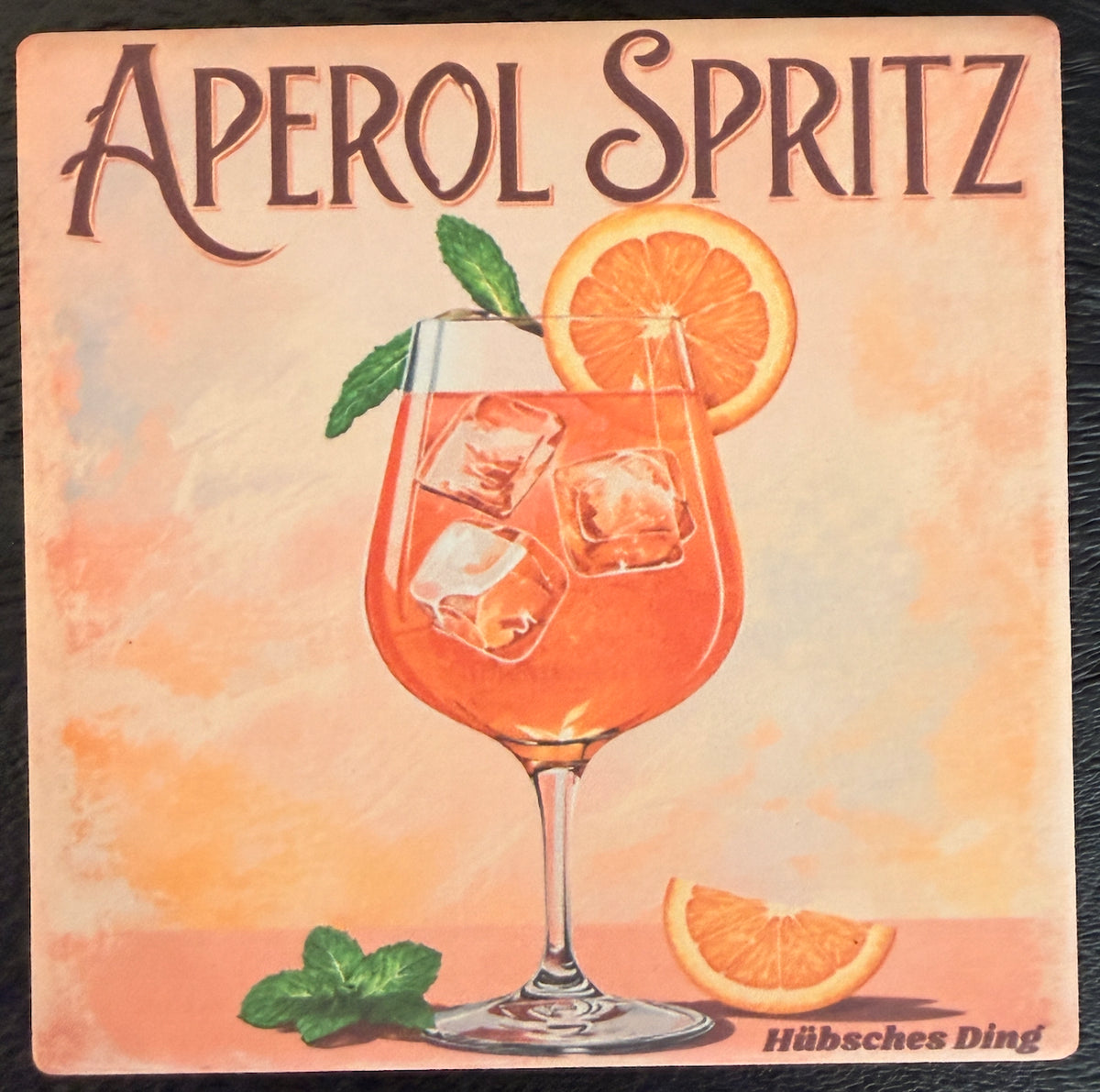 Bedruckte Fliese oder Untersetzer mit einem Aperol Spritz im Weinglas, Eiswürfeln, Orangenscheiben und Minze vor einem warmen Aquarellhintergrund mit dem Schriftzug „Aperol Spritz“.