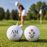 Personalisierte weiße Golfbälle mit Initialen AM und rosafarbenem Blumenmotiv auf einem Golfplatz – bedruckt von Hübsches Ding.