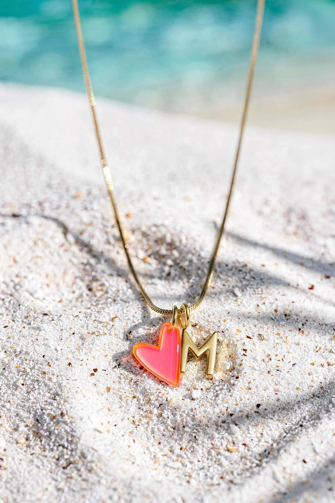 Goldene Halskette mit Buchstaben- und Motiv-Charms von Little Jewellery Stories auf blauem Hintergrund am Strand