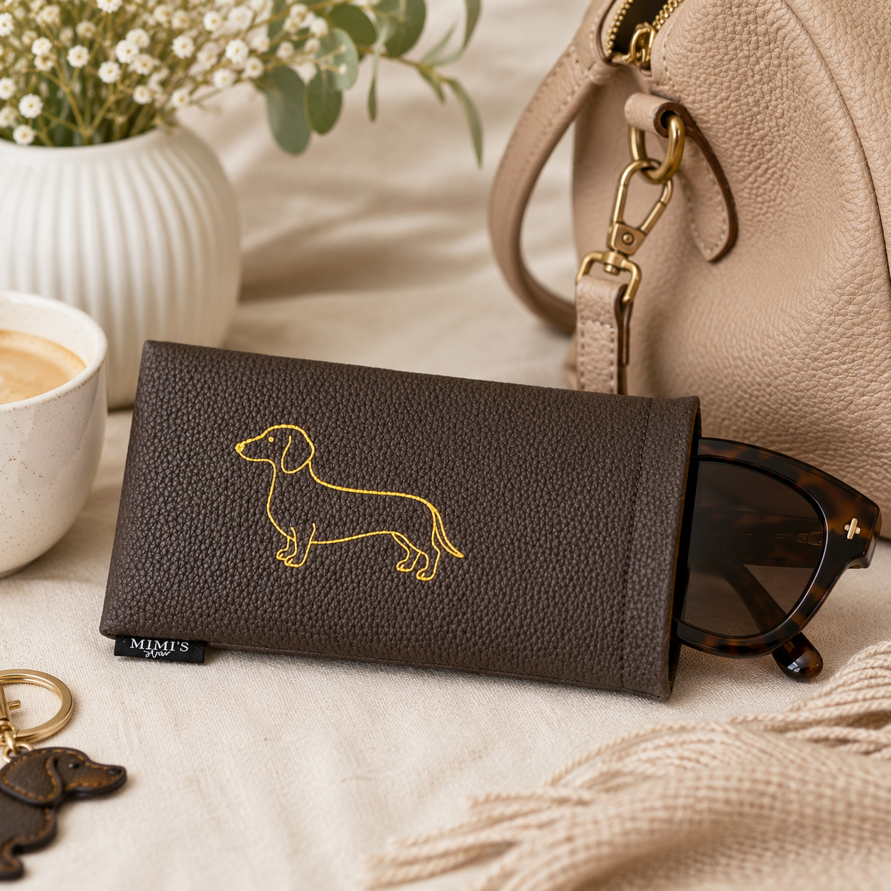 Braunes Ella Brillenetui von Mimi’s Glow mit goldenem Dackel-Motiv, Sonnenbrille, Handtasche und Kaffee in einer warmen Lifestyle-Szene.